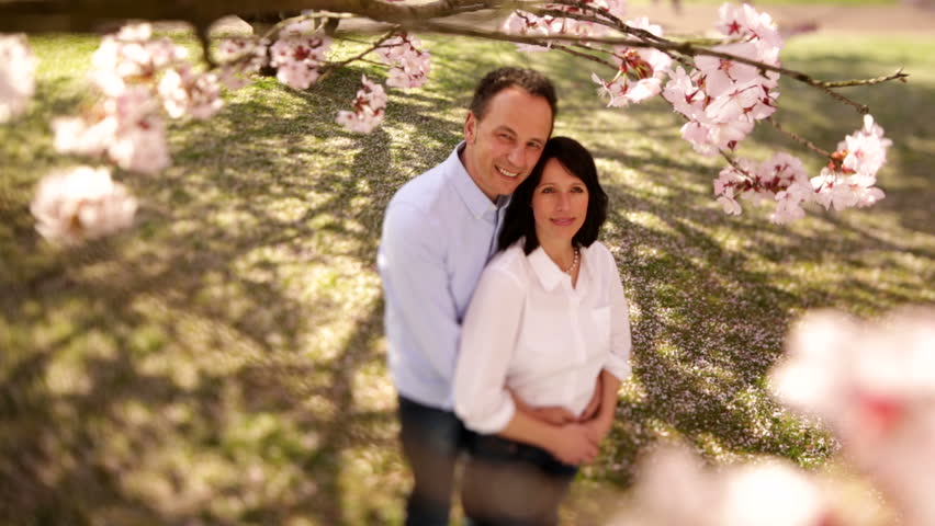 Mature Couple Standing Under Cherry Tree First Looking At Camera Than At Each Other For A Kiss