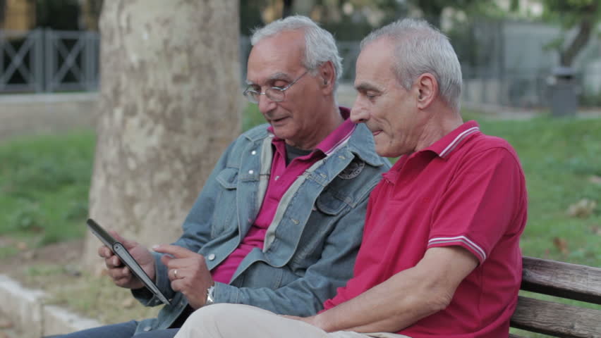 Old Man Teaching His Elderly Friends To Use A New Tablet PC In A Park