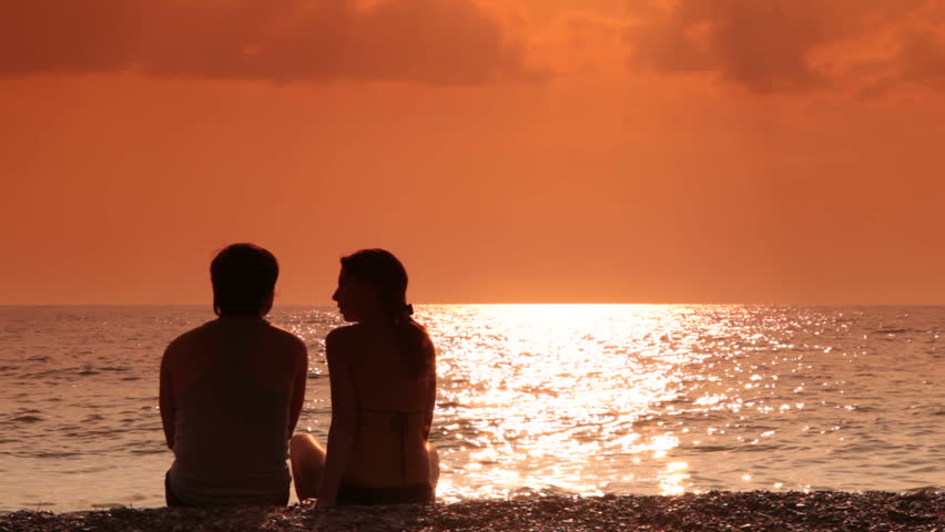 A Couple Sit On The Beach Watching The Sun Set Over The Ocean Stock ...