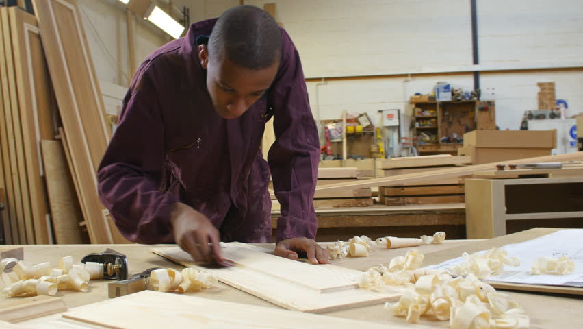 Carpenter Supervising Male Apprentice Sanding Piece Of Wood In ...